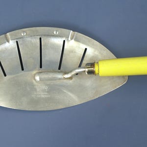 May include: A silver-colored metal hand tool with a curved, fan-shaped blade featuring five vertical slots and a small circular hole on the left side. The tool has a cylindrical, bright yellow handle attached to a central metal shank.