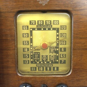 Puede incluir: Primer plano de una radio vintage Emerson con una esfera de color dorado. La esfera tiene una aguja negra que apunta a la marca de 130 kilociclos. La esfera tambi&eacute;n tiene un bot&oacute;n rojo y marcas para diferentes estaciones de radio, incluyendo Francia, EE. UU., Espa&ntilde;a, Alemania, Italia y Jap&oacute;n. La esfera tambi&eacute;n tiene marcas para megaciclos, con la aguja apuntando a la marca de 9 megaciclos. La radio tiene dos botones negros en la parte inferior, uno etiquetado como "TONE".
