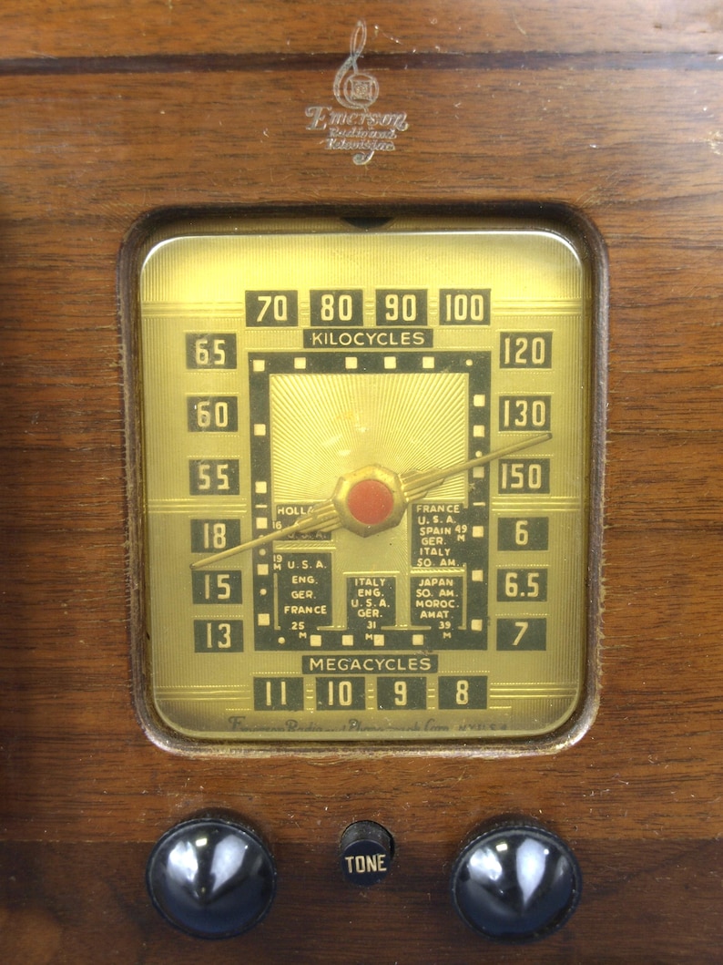 Puede incluir: Primer plano de una radio vintage Emerson con una esfera de color dorado. La esfera tiene una aguja negra que apunta a la marca de 130 kilociclos. La esfera tambi&eacute;n tiene un bot&oacute;n rojo y marcas para diferentes estaciones de radio, incluyendo Francia, EE. UU., Espa&ntilde;a, Alemania, Italia y Jap&oacute;n. La esfera tambi&eacute;n tiene marcas para megaciclos, con la aguja apuntando a la marca de 9 megaciclos. La radio tiene dos botones negros en la parte inferior, uno etiquetado como "TONE".