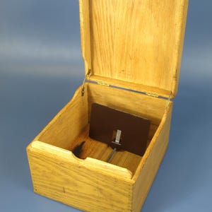May include: An open, wooden box with a hinged lid. The box is made of light-colored wood and has a dark brown rectangular piece inside with a metal rod attached. The box is sitting on a blue surface.