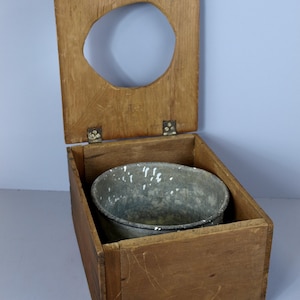 Op de afbeelding: Een houten toiletbril met een metalen emmer erin. De bril is open en de emmer is zichtbaar. Het hout is lichtbruin en de emmer is donkergrijs.