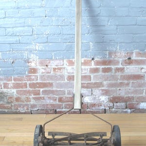 Op de afbeelding: Een antieke duwgrasmaaier met een houten handvat en een metalen snijmechanisme. De maaier heeft twee wielen en staat tegen een bakstenen muur geschilderd met blauwe verf.