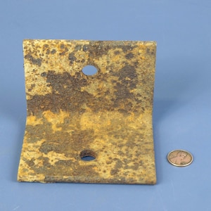 May include: A heavy-duty L-shaped metal bracket with a thick layer of brown and orange rust covering its entire surface. The bracket features two circular holes, one on each side. A copper-colored penny is placed next to it for scale reference.