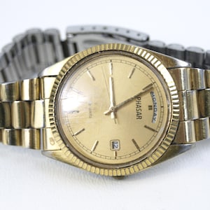 Peut inclure: Une montre analogique dorée avec un cadran doré et un affichage de la date à 3 heures. La montre a une lunette dorée et un bracelet en métal bicolore or et argent. Le cadran de la montre indique "PHASAR" et "MONDAY".