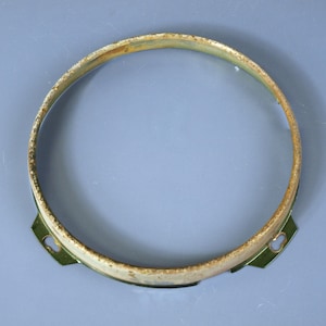 May include: A circular metal headlight trim ring featuring a weathered, rusted finish along the top edge. The component includes two small, flat mounting tabs with oval-shaped holes extending from the bottom sides of the frame.