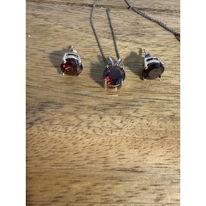 Könnte beinhalten: Ein Schmuckset bestehend aus einer silberfarbenen Halskette mit einem runden, tiefroten facettierten Edelsteinanhänger und zwei passenden runden, tiefroten facettierten Edelsteinohrsteckern, jeweils in einer silberfarbenen Krappenfassung.