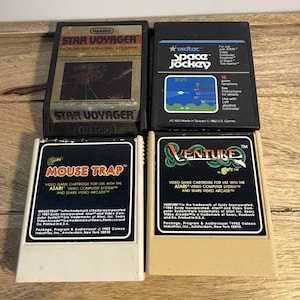 以下が含まれることがあります： Atariシステム用の長方形のビデオゲームカートリッジ4点。左上はStar Voyager、右上はSpace Jockey、左下はMouse Trap、右下はVenture。各カートリッジにはタイトルとテキストが記載されたラベルが貼付されています。