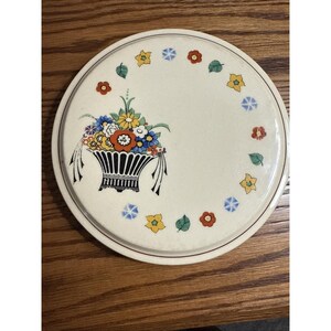 May include: A round, off-white ceramic trivet with a floral basket design. The basket is black with a colorful arrangement of flowers. The border features small flowers and leaves in red, yellow, blue, and green. A thin, dark red line borders the trivet.
