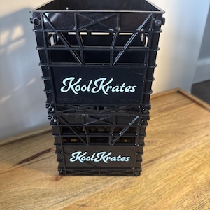 May include: Two black plastic Kool Krate storage crates stacked on top of each other. Each crate has the words "Kool Krate" in white script. The crates are on a wooden surface.