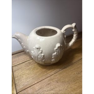 Könnte beinhalten: Cremefarbene Keramik-Teekanne mit rundem Korpus und gebogenem Ausguss. Die Teekanne hat geprägte Blattmotive und einen dekorativen Griff. Die Teekanne steht auf einer Holzoberfläche.