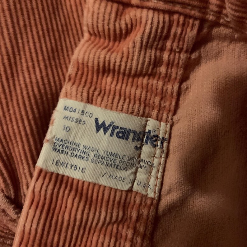 K&ouml;nnte beinhalten: Nahaufnahme einer Hose aus korallenfarbenem Cord. Das Wrangler-Etikett ist sichtbar, mit Text, der die Gr&ouml;&szlig;e und Pflegehinweise angibt. Die Hose hat vertikale Cordrippen.