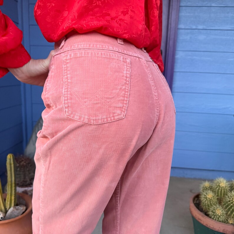 K&ouml;nnte beinhalten: Nahaufnahme einer rosa Cordhose mit Ges&auml;&szlig;taschen. Die Hose hat einen hellen Rosaton und vertikale Cordlinien. Oben im Bild ist eine rote Bluse zu sehen.