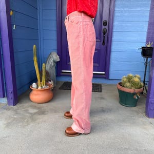 K&ouml;nnte beinhalten: Hellrosa Cordhose mit klassischem F&uuml;nf-Taschen-Design. Die Hose hat einen weiten Beinverlauf und ist am Saum umgeschlagen. Die Person tr&auml;gt braune Lederschuhe. Der Hintergrund umfasst eine blaue T&uuml;r und einen Kaktus im Topf.