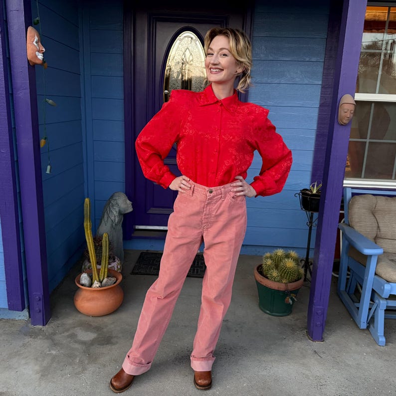 K&ouml;nnte beinhalten: Eine Person tr&auml;gt eine rote Langarmbluse und eine rosa Hose vor einer blauen T&uuml;r. Die Bluse hat ein Blumenmuster und Schulterpolster. Die Hose ist hellrosa. Braune Lederstiefel vervollst&auml;ndigen das Outfit.