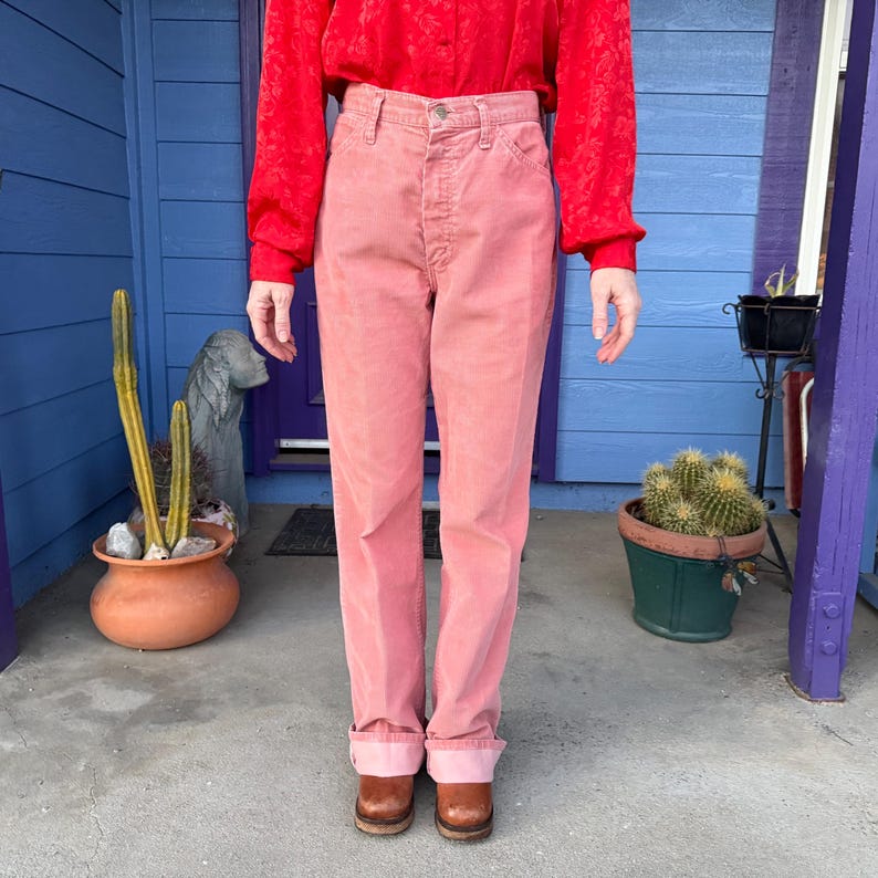 K&ouml;nnte beinhalten: Rosa Cordhose mit umgeschlagenem Saum. Die Hose wird mit braunen Lederschuhen und einem roten Langarmshirt getragen. Der Hintergrund umfasst eine blaue T&uuml;r und Topfpflanzen.