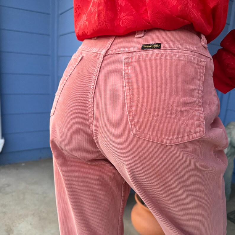 K&ouml;nnte beinhalten: Rosa Cordhose mit einem Wrangler-Etikett auf der Ges&auml;&szlig;tasche. Die Hose hat eine hohe Taille und ein weites Bein. Der Stoff hat vertikale Rippen. Die Hose hat eine ged&auml;mpfte rosa Farbe.