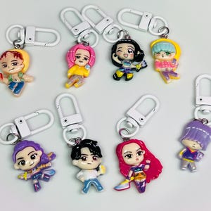 May include: A collection of colorful character keychains with white metal clasps. Each keychain features a miniature figurine with unique hairstyles and outfits. The keychains are in various colors, including pink, yellow, blue, and purple.