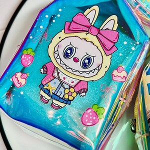 K&ouml;nnte beinhalten: Eine transparente, schillernde Tasche mit einem Cartoon-Hasen-Design. Der Hase tr&auml;gt eine rosa Schleife, Jacke und Latzhose. Erdbeeren und Cupcakes verzieren den blauen Hintergrund. Die Tasche hat einen Rei&szlig;verschluss.