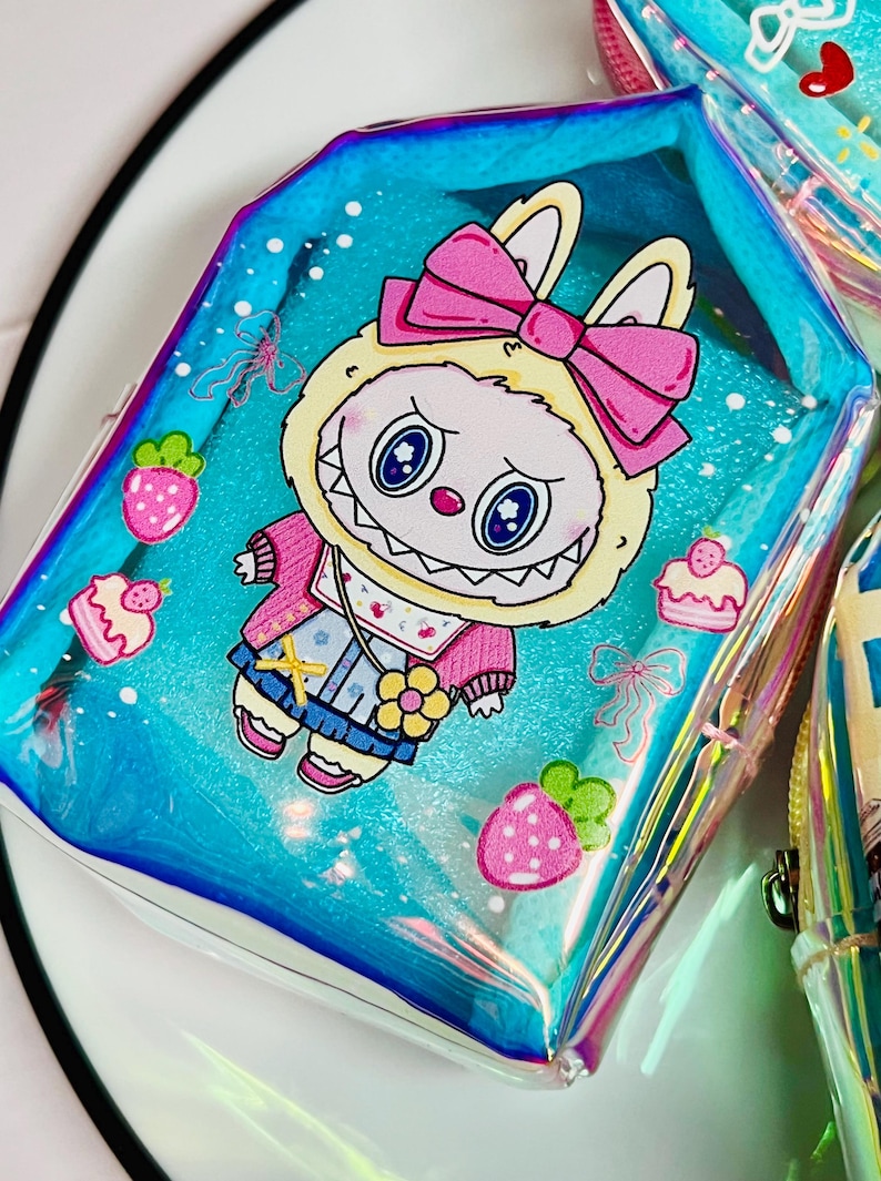 K&ouml;nnte beinhalten: Eine transparente, schillernde Tasche mit einem Cartoon-Hasen-Design. Der Hase tr&auml;gt eine rosa Schleife, Jacke und Latzhose. Erdbeeren und Cupcakes verzieren den blauen Hintergrund. Die Tasche hat einen Rei&szlig;verschluss.