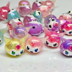 May include: A collection of iridescent pig-shaped beads in various colors, including pink, yellow, blue, and purple. Each bead features a pig face with small ears and a button nose. The beads have a glossy finish and are scattered across a white surface.