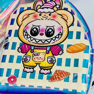 K&ouml;nnte beinhalten: Ein klarer, schillernder Rucksack mit einem Cartoon-B&auml;r-Design. Der B&auml;r tr&auml;gt einen gelben Overall mit einem B&auml;rengesicht. Der Rucksack hat einen blau-wei&szlig; gestreiften Hintergrund mit Bildern von Donut, Croissant und Brot.