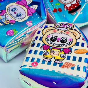 K&ouml;nnte beinhalten: Drei schillernde Kosmetiktaschen mit Cartoon-Charakter-Designs. Eine Tasche zeigt einen B&auml;ren in Latzhosen, eine andere ein H&auml;schen mit einer Schleife und die dritte ein H&auml;schen in einem roten gepunkteten Kleid. Die Taschen haben einen holografischen Glanz.