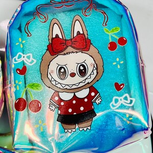K&ouml;nnte beinhalten: Ein klarer, schillernder blauer Rucksack mit einem Cartoon-Hasen-Design. Der Hase tr&auml;gt eine rote Schleife, ein rot-wei&szlig; gepunktetes Kleid und einen schwarzen Rock. Kirschen und Herzen verzieren den Rucksack.