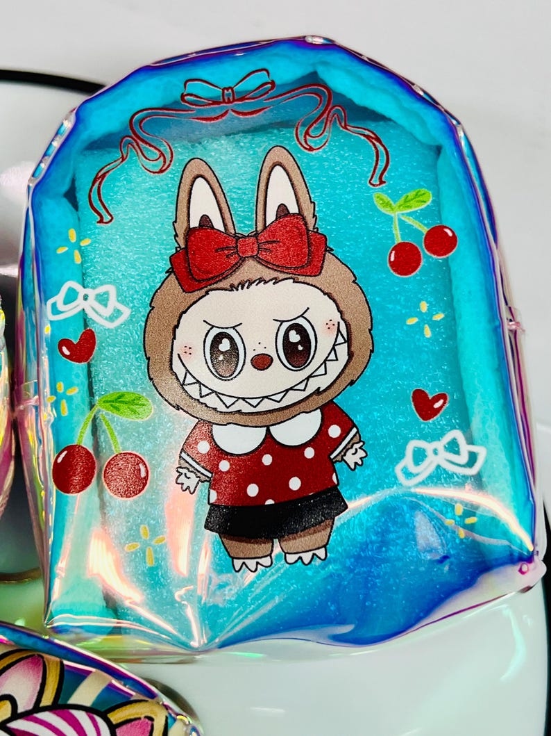 K&ouml;nnte beinhalten: Ein klarer, schillernder blauer Rucksack mit einem Cartoon-Hasen-Design. Der Hase tr&auml;gt eine rote Schleife, ein rot-wei&szlig; gepunktetes Kleid und einen schwarzen Rock. Kirschen und Herzen verzieren den Rucksack.