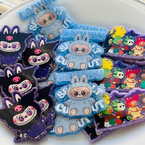 May include: A collection of colorful, cartoonish character pins and barrettes. The pins feature a variety of designs, including a bunny in a blue jumpsuit, and a black cat-like creature. The barrettes have the text "LA BU" on them.