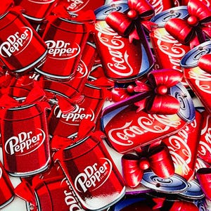 May include: A collection of red and white Dr. Pepper and Coca-Cola can-shaped ornaments, some with red bows. The Dr. Pepper cans have the brand name and logo. The Coca-Cola cans have the brand name in white script. The ornaments are on a white background.