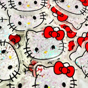 May include: A collection of clear, glitter-filled Hello Kitty face-shaped acrylic charms. Each charm features a red bow, black eyes, whiskers, and a yellow nose. The charms are outlined in black and filled with iridescent glitter.
