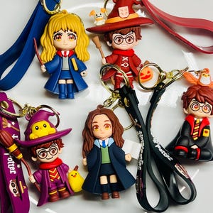 May include: A collection of character keychains featuring figures in school uniforms and wizard attire. The keychains have gold-colored metal rings and colorful wrist straps. The figures are in various poses, some holding wands or books.