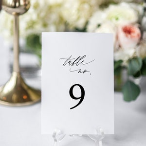 May include: White card with black calligraphy that reads "table no. 9" on a clear acrylic stand. The card is sitting on a white tablecloth.