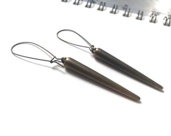 Può includere: Un paio di orecchini allungati color bronzo con un design conico a punta. Gli orecchini hanno un semplice gancio di filo per orecchie forate. Gli orecchini sono su uno sfondo bianco.