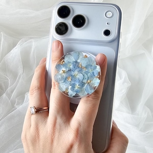 May include: A circular phone grip featuring a clear resin dome embedded with light blue hydrangea petals and small gold leaf flakes, attached to the back of a light gray smartphone case with a triple camera lens array on the upper left corner.