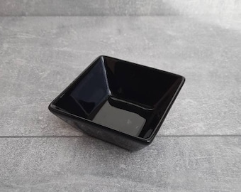 May include: A small, square, black ceramic bowl. The bowl has a glossy finish and is set against a gray background. The bowl's shape is geometric, with angled sides and a flat bottom. It is likely used for serving dips or small portions.