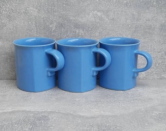May include: Three light blue ceramic mugs with angular sides and rounded handles are arranged in a row. The mugs have a simple, modern design and are set against a gray background.