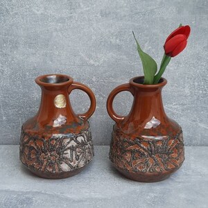 May include: Two brown ceramic vases with handles, featuring a textured floral design on the lower half. One vase holds a red tulip. The vases have a glossy finish and a vintage aesthetic, suitable for home decor.