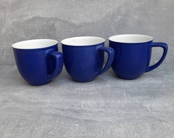 May include: Three cobalt blue ceramic mugs with white interiors are arranged side by side. Each mug has a rounded body, a curved handle, and a smooth, glossy finish. The mugs are set against a gray background.