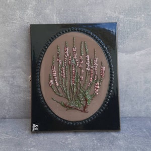 Könnte beinhalten: Quadratische, schwarze Keramikfliese mit einem erhabenen ovalen Design mit einer botanischen Illustration. Die Illustration zeigt einen Cluster aus rosa und grünen Blumen auf braunem Hintergrund, umrahmt von einem Perlenrand.