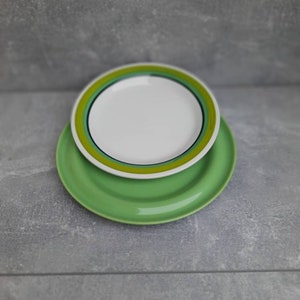 May include: Two ceramic plates. The bottom plate is a solid light green. The top plate is white with a green and yellow border. The plates are stacked on top of each other.