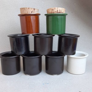 May include: A collection of ceramic jars in various colors, including brown, green, black, and white. Each jar has a cylindrical shape and a cork lid. The jars are stacked, showcasing their different hues and textures.