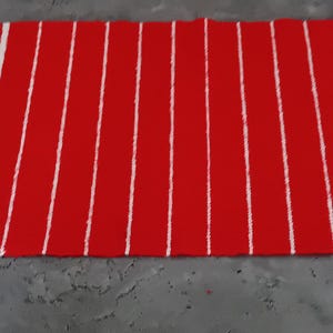 May include: A rectangular red and white striped rug. The rug has a solid red border on the top and bottom, with vertical white stripes alternating with red stripes in the center. The rug is made of a woven material.