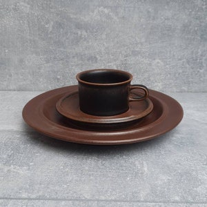 May include: A brown ceramic coffee cup and saucer set. The cup sits on a small saucer, which is on a larger plate. The set is a solid brown color and appears to be made of stoneware. The set is designed for serving coffee or tea.