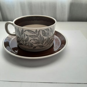 May include: A vintage teacup and saucer set. The cup and saucer are brown and white with a floral pattern. The cup has a brown interior and a grey handle. The saucer is round with a brown rim.