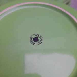 May include: Close-up of a stack of green ceramic plates. The plates have a light green color with a pink rim. The bottom of the plate has a circular logo that says "HOGANAS KERAMIK".