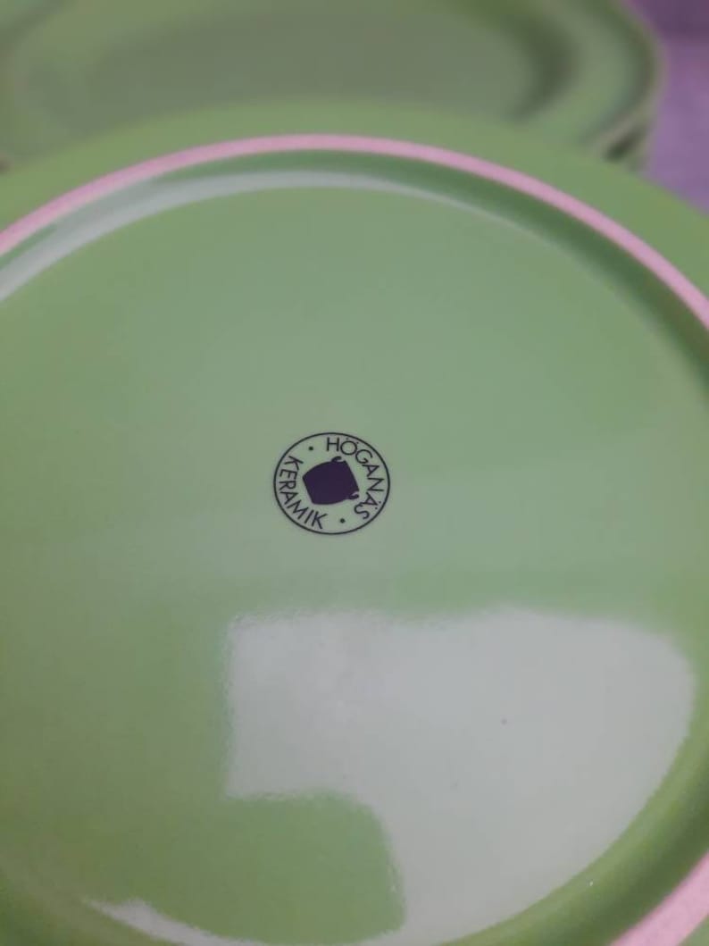 May include: Close-up of a stack of green ceramic plates. The plates have a light green color with a pink rim. The bottom of the plate has a circular logo that says "HOGANAS KERAMIK".