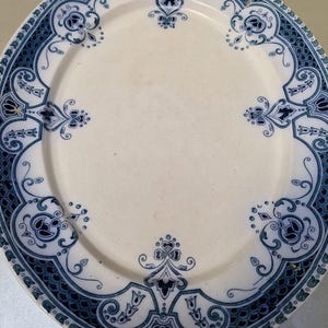Pode incluir: Um prato redondo de cerâmica com um centro de cor creme e uma borda larga com um padrão intrincado em azul escuro. O design da borda inclui motivos florais repetitivos, volutas e uma textura semelhante a escamas na borda externa.