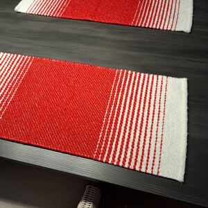 May include: Two rectangular red and white woven table runners. The runners have a gradient design, transitioning from solid red to white stripes. The runners are placed on a dark wooden table.