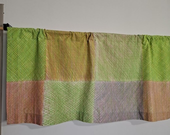 May include: A valance curtain with a patchwork design. The curtain features squares of green, orange, and purple fabric. The fabric has a textured, woven appearance. The curtain is hanging on a wooden rod against a white wall.